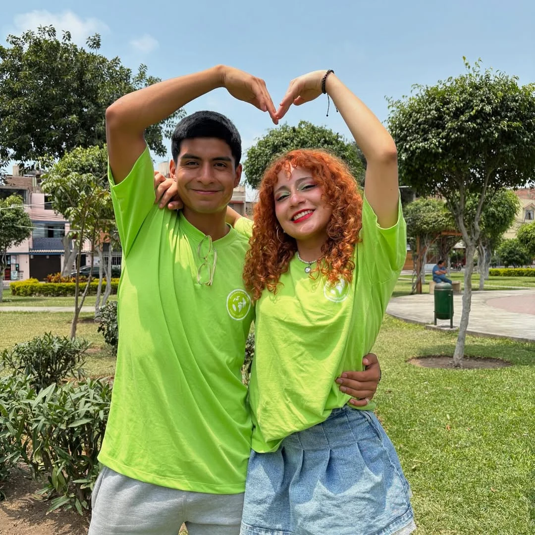 Dos voluntarios con polos lima de BUC forman un corazón con los brazos en un parque